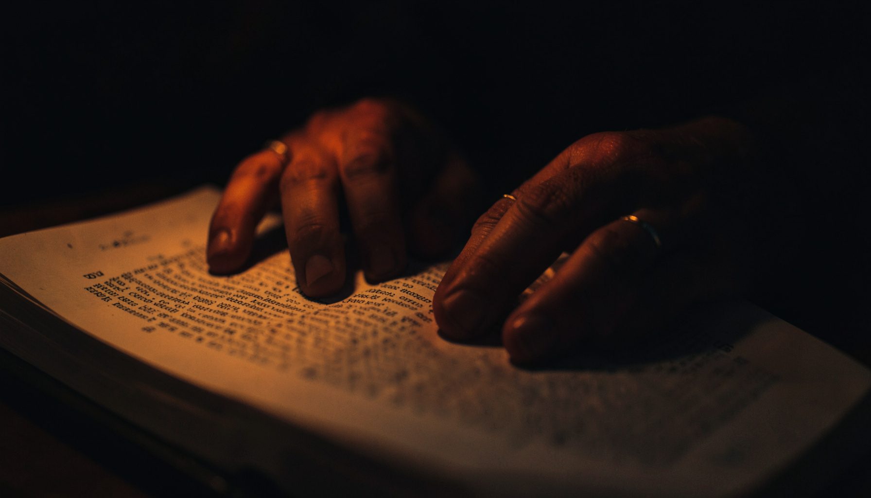 Dark, low-key photograph of hands gently resting on an open book of scripture, with soft warm light illuminating the pages.