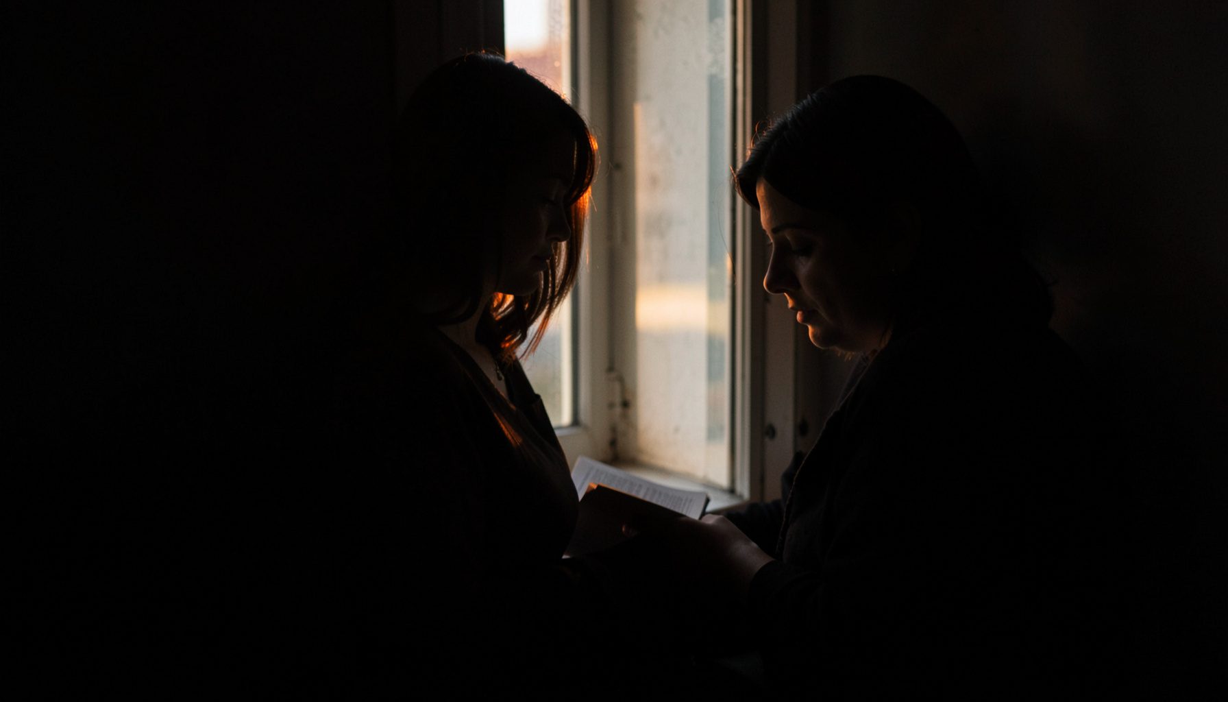 Dark, low-key photograph of a woman reading near a window, gentle morning light, contemplative mood.