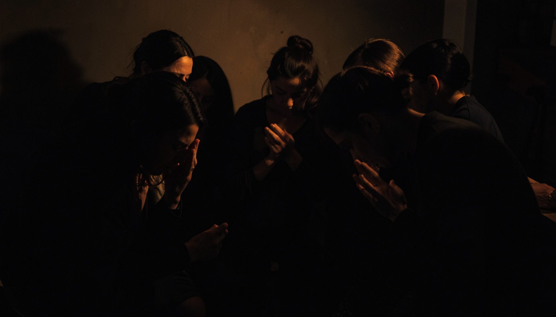 Dark, low-key photograph of several women gathered in a circle, heads bowed in prayer, soft amber light.