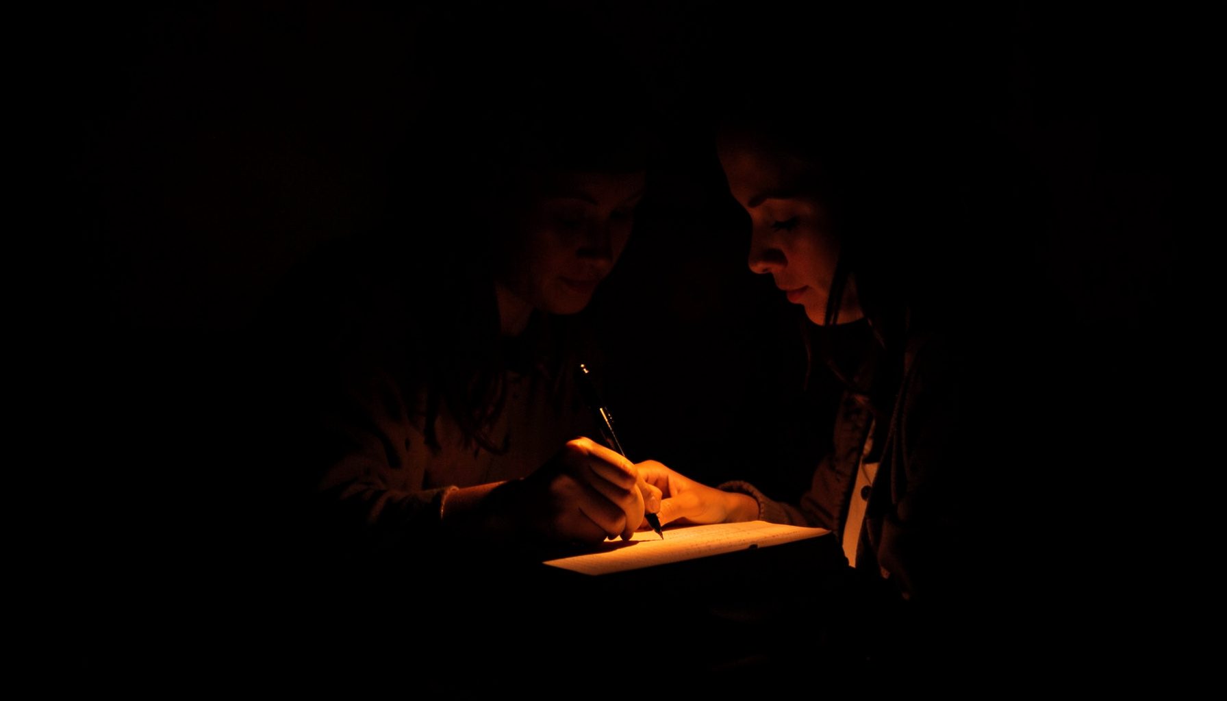 Dark, low-key photograph of a woman writing in a journal, warm lamplight casting deep shadows.