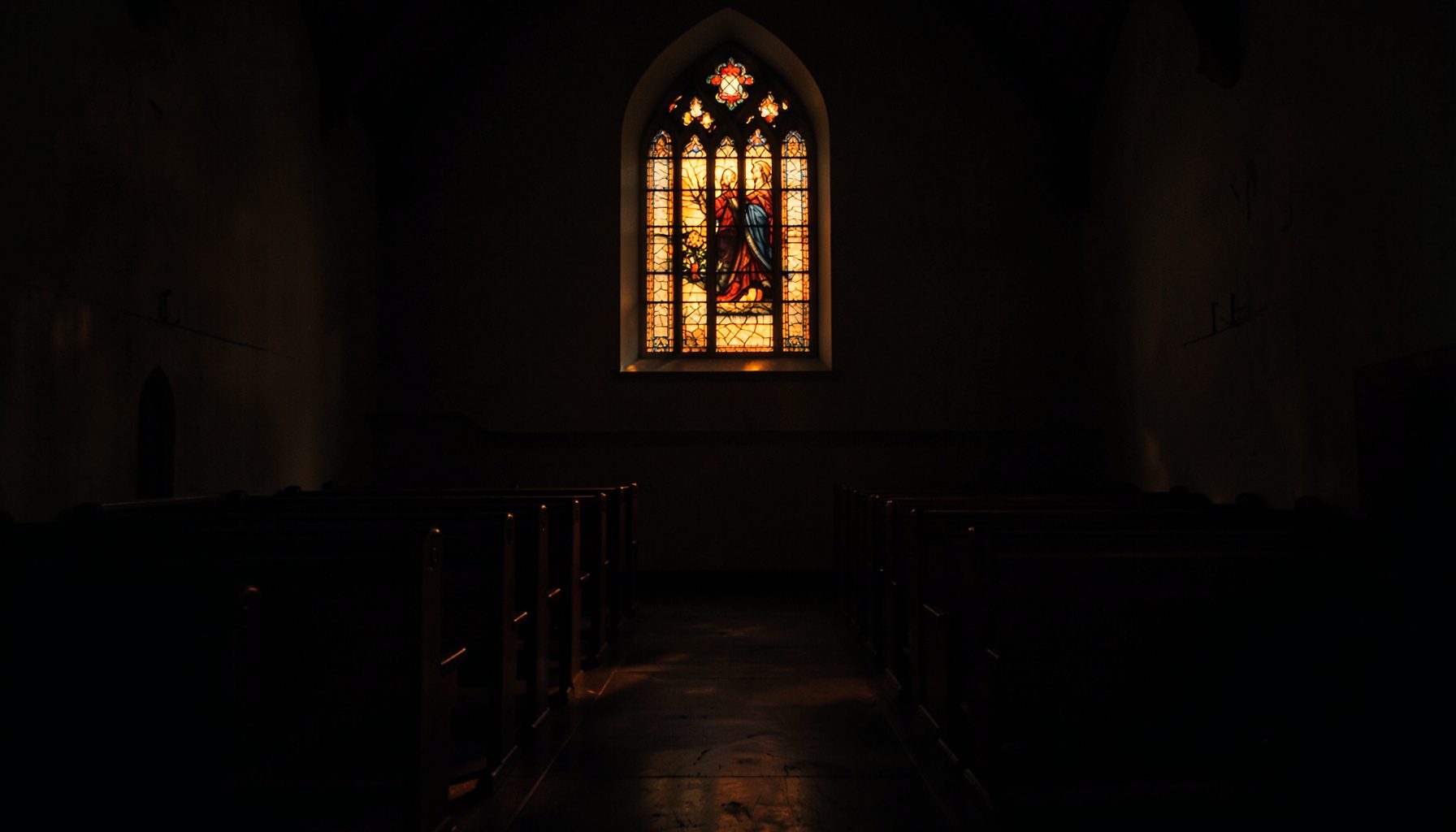 Dark, low-key photograph of a quiet chapel interior with wooden pews and soft amber light filtering through a stained-glass window, creating a reverent and contemplative mood.