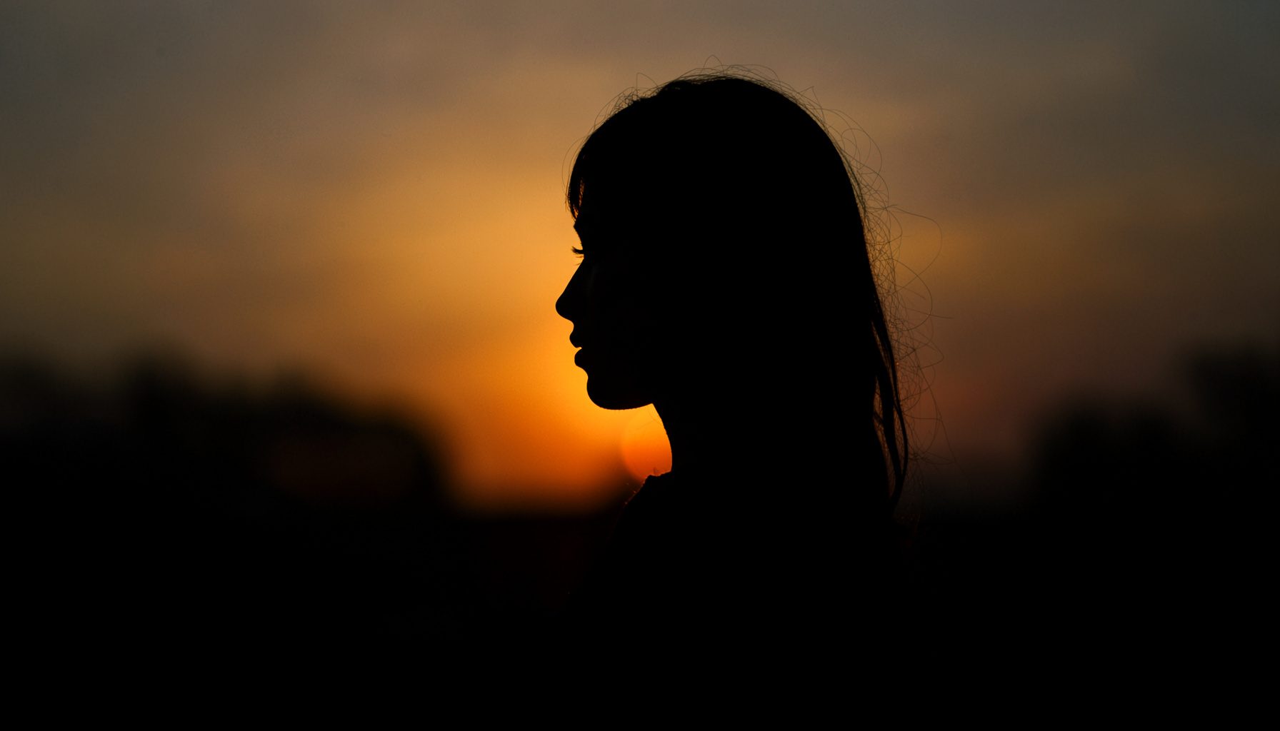 Dark, low-key photograph of a woman's silhouette against a sunset, golden rim light, peaceful atmosphere.