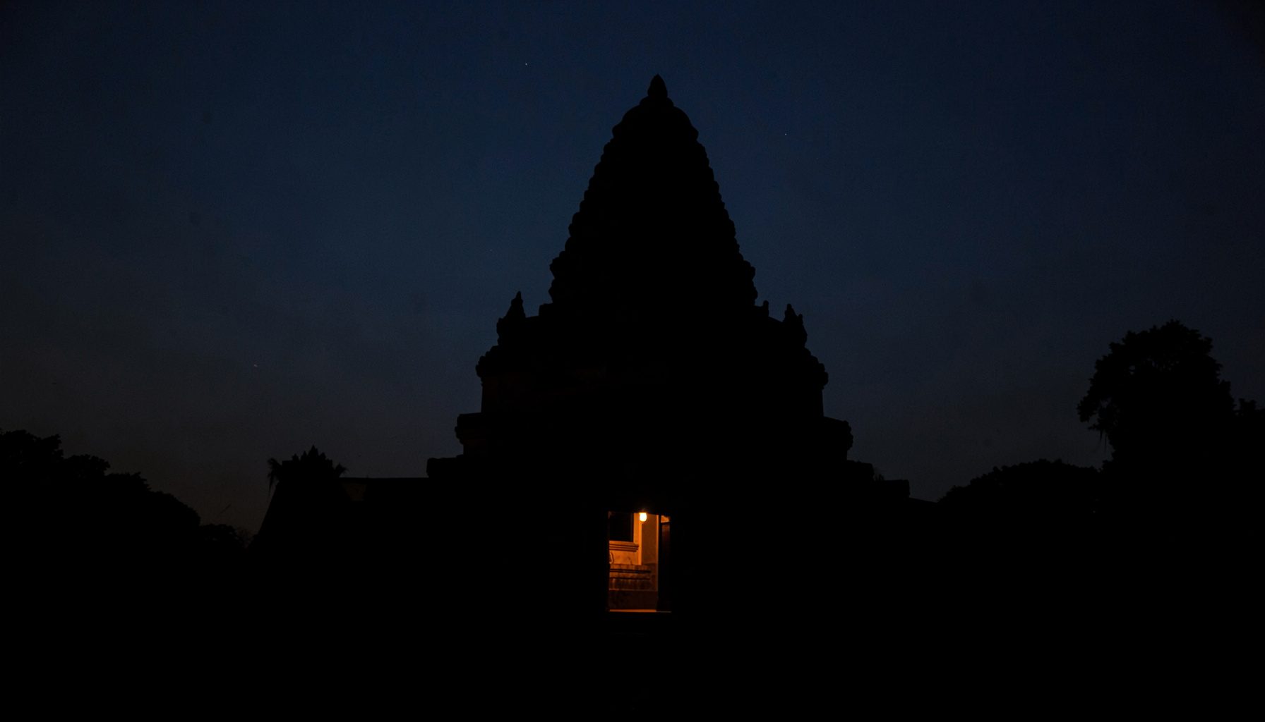 Dark, low-key photograph of a temple silhouette against a deep twilight sky, a single warm light glowing from within.