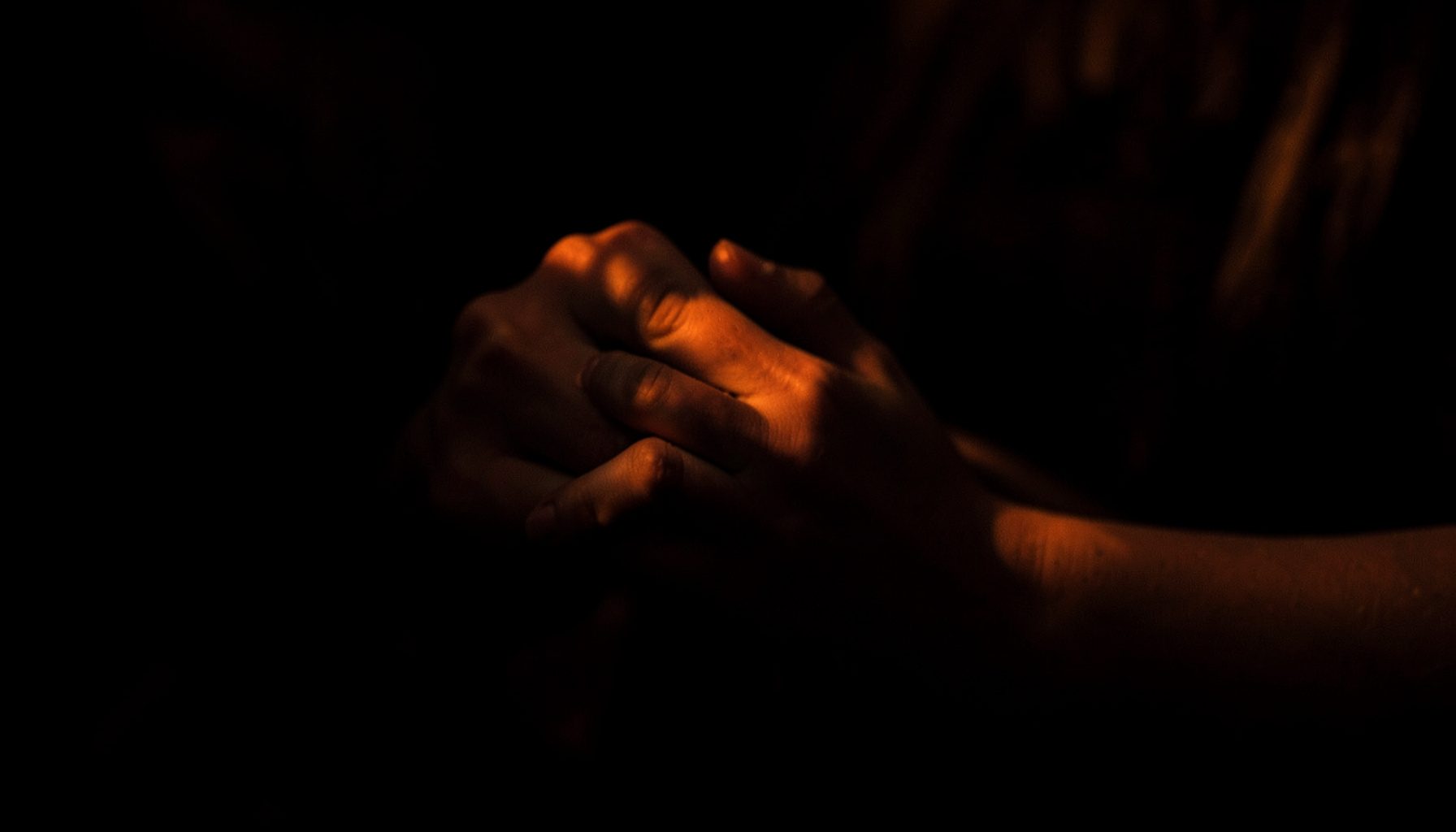 Dark, low-key portrait-style photograph of a woman's hands folded in quiet contemplation, soft amber light.