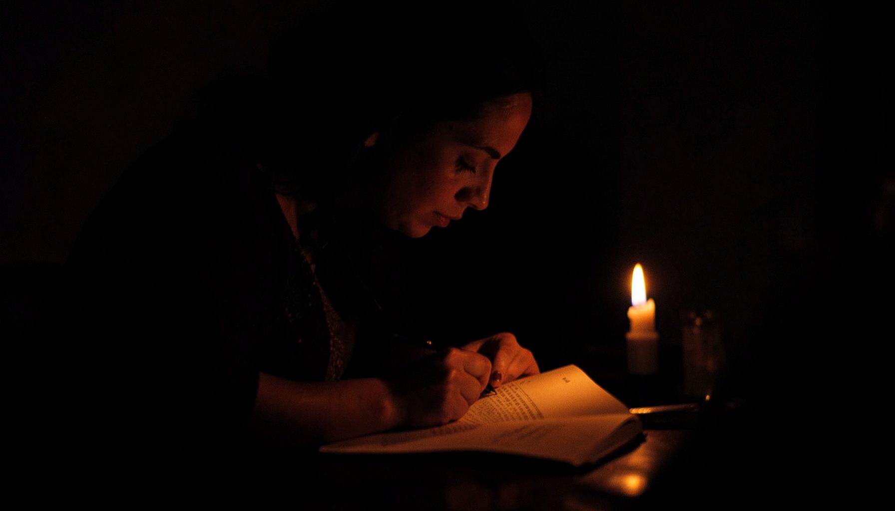 Dark, low-key photograph of a woman writing thoughtfully in a journal by candlelight, deep shadows and warm highlights.